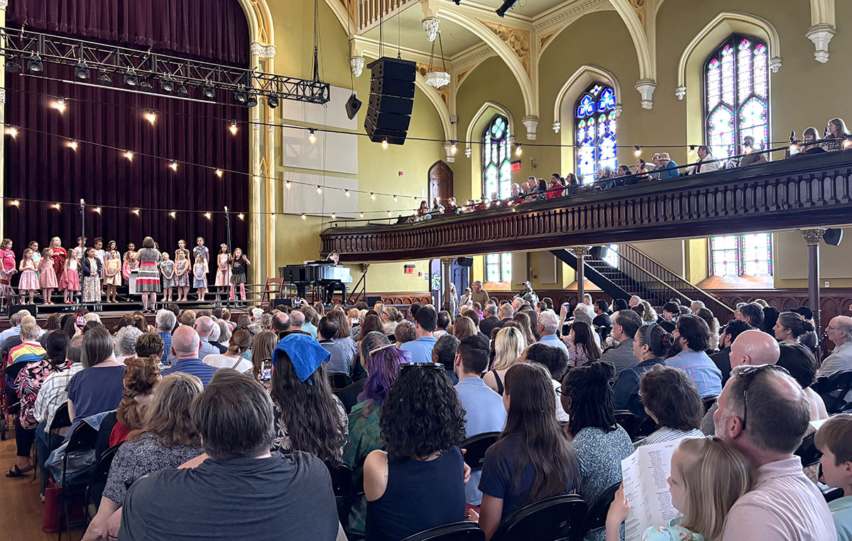 Audience view of Buffalo Girlchoir performing on a church stage with a crowded balcony and tall stained glass windows.